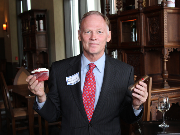 Will McDonald, dressed in a suit, holding a red velvet cupcake in one hand and a cigar in the other.