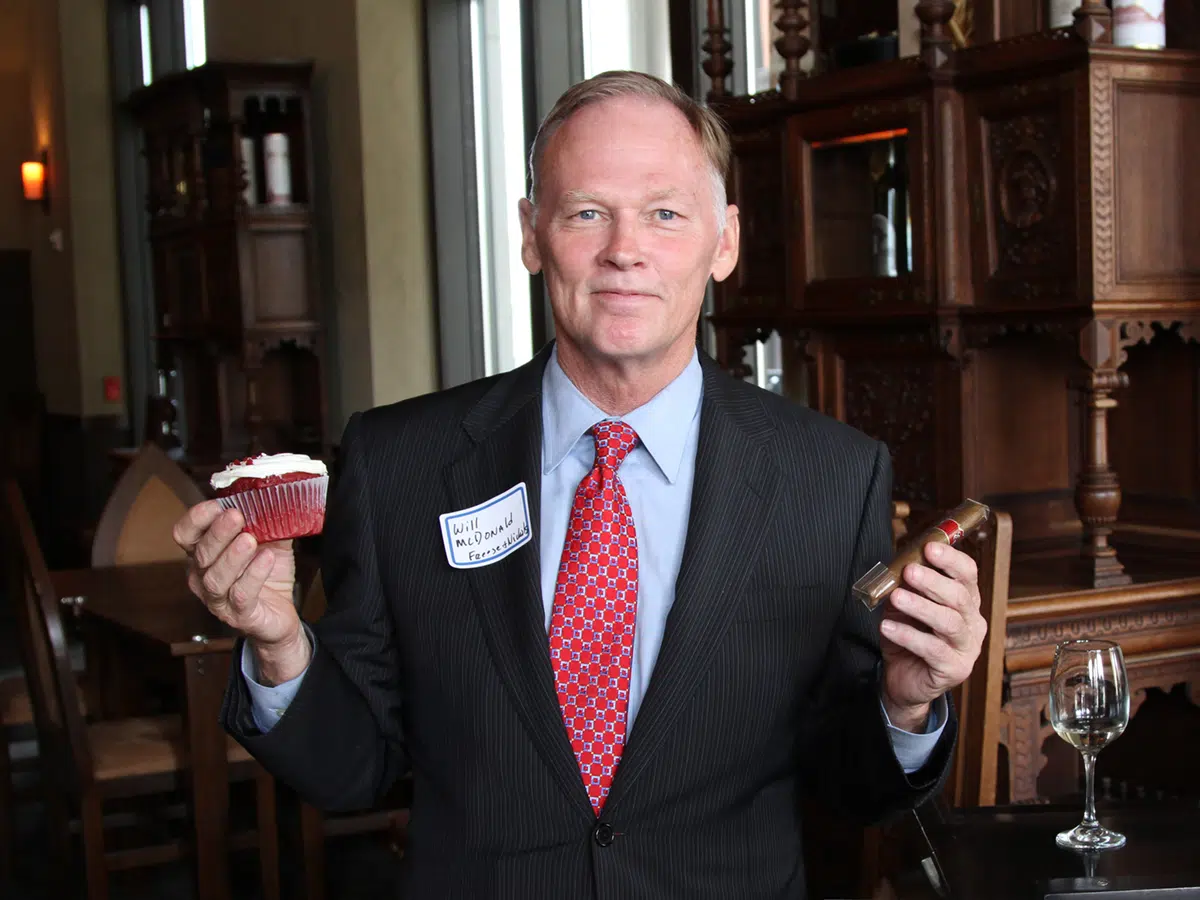 Will McDonald, dressed in a suit, holding a red velvet cupcake in one hand and a cigar in the other.